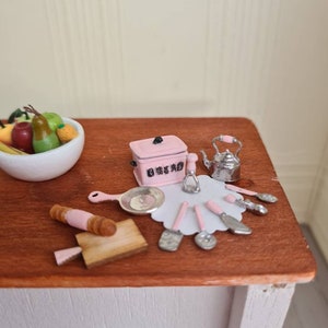 May include: A miniature kitchen set with a pink bread box, a silver teapot, a rolling pin, a wooden cutting board, and a set of silver kitchen utensils on a wooden countertop.