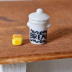 May include: White ceramic bread jar with a black inscription that reads "Bread". A small yellow bread loaf sits next to the jar on a wooden surface.