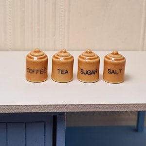 Peut inclure: Quatre petits pots en céramique avec couvercles, étiquetés "Café", "Thé", "Sucre" et "Sel". Les pots sont brun et blanc et sont posés sur une surface blanche.