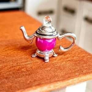 Könnte beinhalten: Eine silberne Miniatur-Teekanne mit einem rosafarbenen Deckel und einem durchsichtigen Glasknopf. Die Teekanne steht auf einem Holztisch.