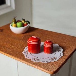May include: Two red ceramic canisters with white trim, sitting on a white doily on a wooden table.