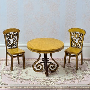 May include: Miniature wooden dining set featuring a round table and two chairs. The table has a dark brown, ornate base and a light brown tabletop. The chairs have decorative backrests. The set is placed on a pink and white floral rug.