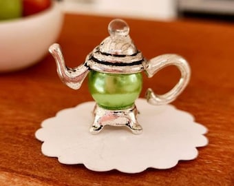 May include: A miniature silver teapot with a green glass body and a white lace doily. The teapot is sitting on a wooden surface.