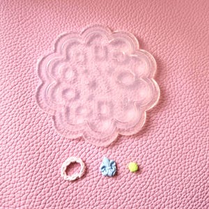 Puede incluir: Un molde transparente en forma de flor con varias formas pequeñas, junto con una goma para el pelo blanca, un objeto azul y una pequeña perla amarilla. Los artículos están dispuestos sobre una superficie rosa texturizada, probablemente para manualidades o joyería.