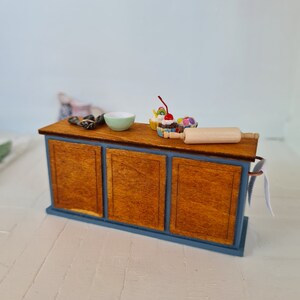 May include: A miniature wooden kitchen counter with a blue base and three drawers. The counter is decorated with a small green bowl, a rolling pin, and three miniature cupcakes with colorful frosting and a red cherry on top.