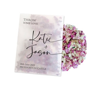 May include: White paper bag with the text "THROW SOME LOVE" and "Katie + Jason" in black script. The date "28th June 2022" and "PECKFORTON CASTLE" are also printed on the bag. The bag is filled with pink and white flower petals.