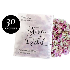 May include: Thirty white paper packets with pink and white flower petals, labeled "Throw Some Love" with the names "Steven + Rachel" and the date "Friday 8th April 2022" and the location "Rothienorman".