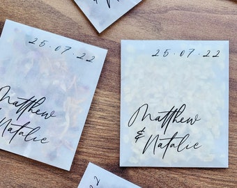May include: Four white paper bags with black calligraphy writing. The bags contain confetti and have the date "25.07.22" and the names "Matthew & Natalie" written on them.