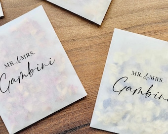 May include: White paper bags with the text "MR. & MRS. Gambini" printed in black. The bags are filled with white and pink confetti.