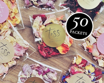 May include: Fifty individual packets of rose petals, each with a gold circle sticker that says "Toss at the Newlyweds". The packets are arranged on a wooden surface.