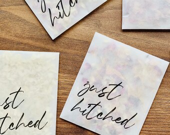 May include: Four white paper bags with the text "just hitched" written in black calligraphy. The bags are filled with confetti and are arranged on a wooden surface.