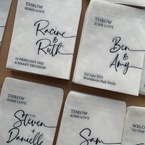 May include: White paper bags with black calligraphy text. The bags have the text "Throw some love" and the names of couples getting married. The text includes the date and location of the wedding.