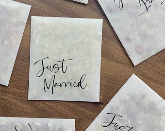 May include: Five white paper bags with the text "Just Married" written in black calligraphy. The bags are filled with confetti and are arranged on a wooden surface.