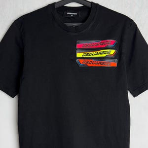 Może przedstawiać: Czarny T-shirt z krótkim rękawem z wielokolorowym nadrukiem słowa "DSQUARED2" powtórzonego cztery razy po lewej stronie klatki piersiowej.