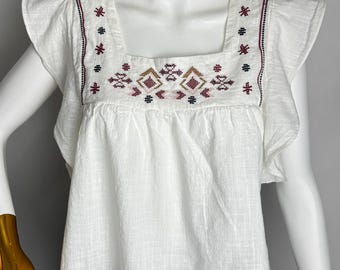 May include: White square-neck top with embroidered details in red, brown, and black. The top features short, ruffled sleeves and a loose fit. The garment is displayed on a mannequin.