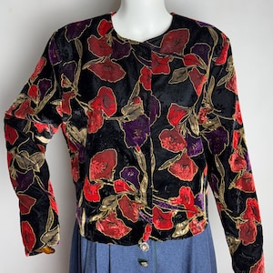 Op de afbeelding: Een zwart fluwelen jasje met een bloemenprint in tinten rood, paars en goud. Het jasje heeft lange mouwen en een knoopsluiting.