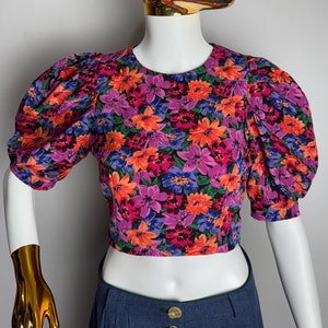 Könnte beinhalten: Eine geblümte Bluse mit Puffärmeln und verkürzter Länge. Die Bluse hat einen schwarzen Hintergrund mit rosa, orangefarbenen und blauen Blumen.