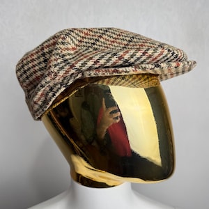 May include: A tweed newsboy cap with a classic plaid pattern in shades of brown, red, and black. The cap is displayed on a gold mannequin head, showcasing its shape and design. This hat is a stylish accessory for various outfits.