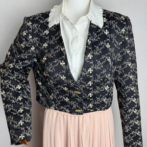 May include: A black cropped blazer with a floral pattern in cream-colored thread. The blazer has long sleeves and gold-colored buttons. It is paired with a white collared shirt and a light pink pleated skirt.