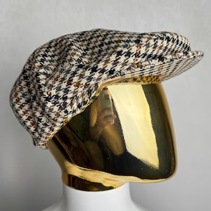 May include: A tweed newsboy cap with a classic houndstooth pattern in black, white, and brown. The cap is displayed on a gold mannequin head, showcasing its shape and style. This hat is a stylish accessory.