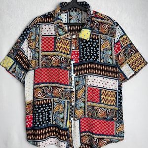 May include: Short-sleeved button-down shirt with a patchwork design. The shirt features a variety of patterns, including paisley, polka dots, and geometric shapes, in a mix of colours such as red, blue, yellow, and brown. The shirt is hanging on a black hanger.