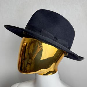 Puede incluir: Un sombrero fedora de color azul marino oscuro con una cinta de grosgrain a juego alrededor de la copa. El sombrero se exhibe sobre una cabeza de maniquí con un rostro dorado pulido y reflectante que muestra un reflejo distorsionado.