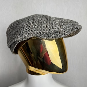 Puede incluir: Gorra de estilo newsboy con estampado de espiga gris y negro. La gorra se exhibe sobre una cabeza de maniquí dorada. La gorra tiene una corona redondeada y una visera corta y rígida. La tela tiene una apariencia texturizada.