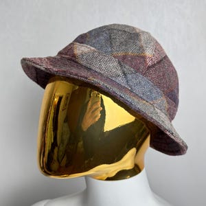 Könnte beinhalten: Ein karierter Tweedhut in Lila-, Blau- und Grautönen. Der Hut hat eine kleine Krempe und wird auf einem goldenen Schaufensterkopf präsentiert. Der Hut ist aus einem strukturierten Stoff gefertigt.