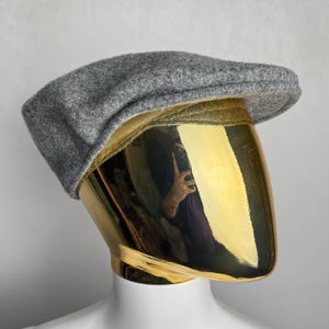 Pode incluir: Bon&eacute; plano cinzento de l&atilde;, com coroa arredondada e aba curta e r&iacute;gida. O chap&eacute;u &eacute; exibido em uma cabe&ccedil;a de manequim dourada sobre um fundo branco. O bon&eacute; &eacute; um estilo cl&aacute;ssico, adequado para v&aacute;rias ocasi&otilde;es.