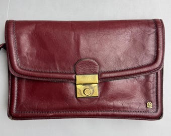 May include: A burgundy leather clutch with a rectangular shape. The clutch features a flap closure secured by a gold-colored metal clasp. The leather appears to be slightly worn, adding to its vintage appeal. A small, circular logo is visible on the lower right corner.