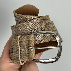 Puede incluir: Un cinturón con estampado de piel de serpiente en tonos beige y plateado con una hebilla plateada. El cinturón de cuero tiene una superficie texturizada. El cinturón está enrollado, revelando el interior de cuero.