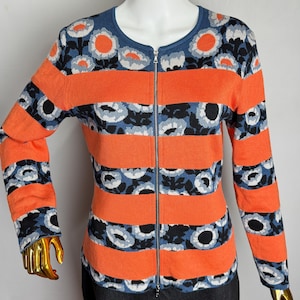 Pode incluir: Um suéter com zíper com estampa floral e listras laranja. O suéter apresenta decote e punhos azuis, com um padrão repetido de flores brancas e pretas sobre um fundo azul. As listras laranja são horizontais.