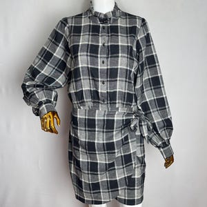 May include: A long-sleeved dress featuring a black and white plaid pattern, a high ruffled collar, and a button-down front. The garment includes a wrap-style skirt with a side tie detail, long sleeves with cuffs, and a structured bodice design.