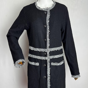 Puede incluir: Abrigo largo negro con cuello redondo y mangas largas. El abrigo presenta un ribete decorativo en tejido tipo tweed blanco y negro a lo largo de los bordes, bolsillos y puños. Tiene botones negros en la parte delantera.