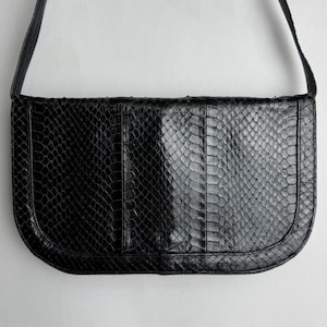 Könnte beinhalten: Schwarze Umhängetasche mit Schlangenmuster. Die Tasche hat eine rechteckige Form mit abgerundeter Unterseite und einen langen, dünnen Riemen. Der Riemen ist oben an der Tasche befestigt. Der Hintergrund ist weiß.
