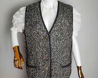 Op de afbeelding: Een zwart-witte tweedvest met een zwarte bies en vier bruine knopen. De vest heeft twee zakken aan de onderkant.