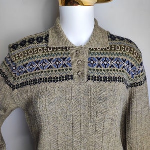 May include: A tan sweater with a collared neckline and three buttons. The yoke features a geometric pattern in blue, green, and black. The sweater has a ribbed texture.
