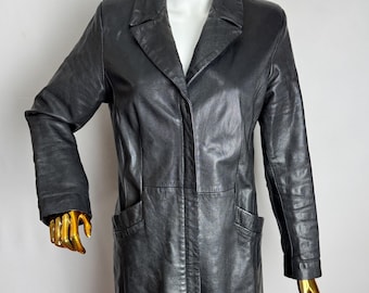 May include: A black leather coat with a notched collar and two front pockets. The coat is knee-length and has long sleeves. The coat is displayed on a mannequin with a gold head and hands.