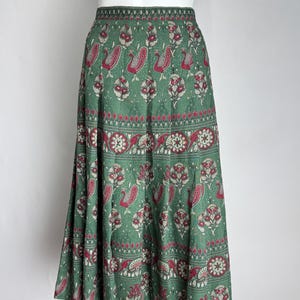 May include: Green skirt with a repeating pattern of red and white floral and peacock designs. The skirt has a wide waistband and a flowing, A-line shape. The fabric appears to be lightweight, suitable for warm weather.