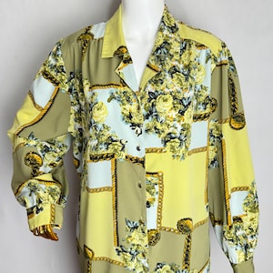 May include: A long-sleeved blouse with a floral and chain pattern. The blouse features a mix of yellow, olive green, and light blue panels. The floral design includes yellow roses. The blouse has a collared neckline and button closure.