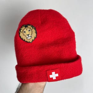 Könnte beinhalten: Eine leuchtend rote Beanie-Mütze mit umgeschlagenem Saum. Sie hat einen gold-braunen Löwenaufnäher an der Seite und ein weißes Quadrat mit rotem Kreuz am Saum. Die Mütze ist aus weichem Strickmaterial gefertigt.