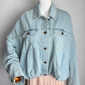 May include: Light blue denim jacket with a classic collar, button-down front, and two chest pockets. The jacket has a slightly oversized fit and features dark brown buttons. The sleeves are cuffed, and the jacket is displayed on a mannequin.