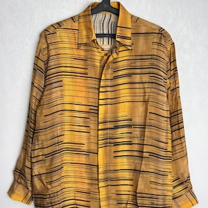 Puede incluir: Una camisa de manga larga con botones y un estampado de rayas amarillas y negras. La camisa está colgada de una percha negra sobre un fondo blanco. El patrón consiste en líneas negras horizontales sobre una base amarilla.