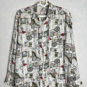 May include: White button-down shirt with a geometric pattern in grey, green, and red. The shirt has long sleeves, a collar, and the brand name "ACCANTO" on the label. The shirt is hanging on a white hanger.