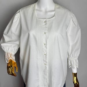 May include: A white blouse with a square neckline and long sleeves. The blouse features pleats down the front and lace detailing at the cuffs. The sleeves are gathered at the shoulders and cuffs. The blouse is buttoned down the front.