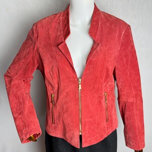 Puede incluir: Chaqueta de ante color coral con cuello alzado y cremallera dorada completa. La chaqueta tiene dos bolsillos con cremallera y mangas largas. La chaqueta se exhibe en un maniqu&iacute;.