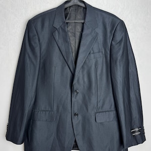 Puede incluir: Un blazer gris oscuro de dos botones con un sutil patrón de rayas verticales, colgado de una percha negra. La chaqueta tiene dos bolsillos delanteros y una solapa con muescas. El puño de la manga tiene cuatro botones. Una etiqueta es visible en la manga.