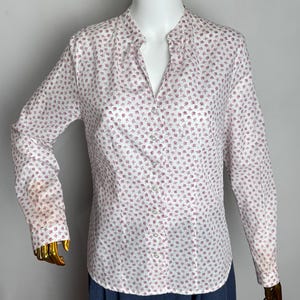 Puede incluir: Camisa blanca de manga larga con un pequeño estampado floral rosa repetido. La camisa tiene cuello mandarín y cierre de botones. Las mangas son largas y la camisa es de tejido ligero. Se combina con una falda azul.