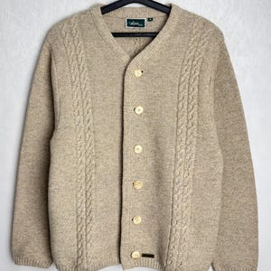 May include: A tan cardigan sweater with a V-neck and button closure. The sweater features a cable knit design along the front and sleeves. The label reads "Alpin" in green. The sweater is hanging on a black hanger.