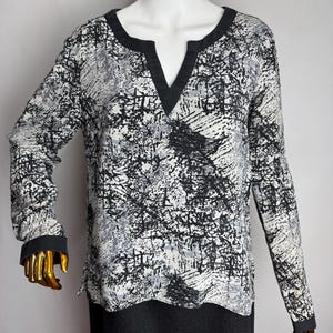 Könnte beinhalten: Eine langärmlige Bluse mit einem abstrakten Schwarz-Weiß-Muster. Die Bluse hat einen V-Ausschnitt und schwarze Verzierungen an Kragen und Manschetten. Der Stoff hat ein strukturiertes Aussehen, und der Stil ist lässig.
